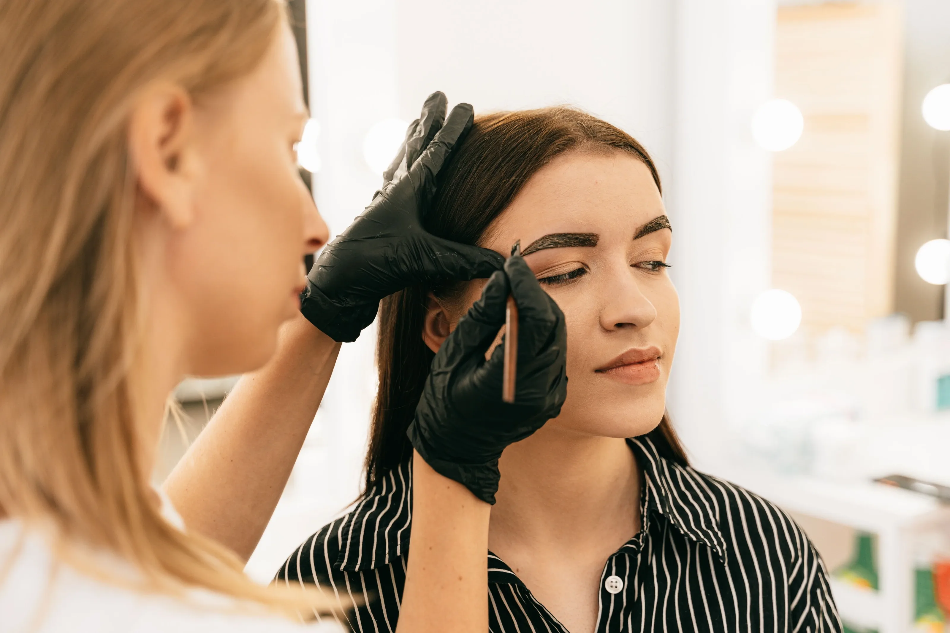 Brow artist in gloves checking a client's eyebrows in a salon with mirror lights