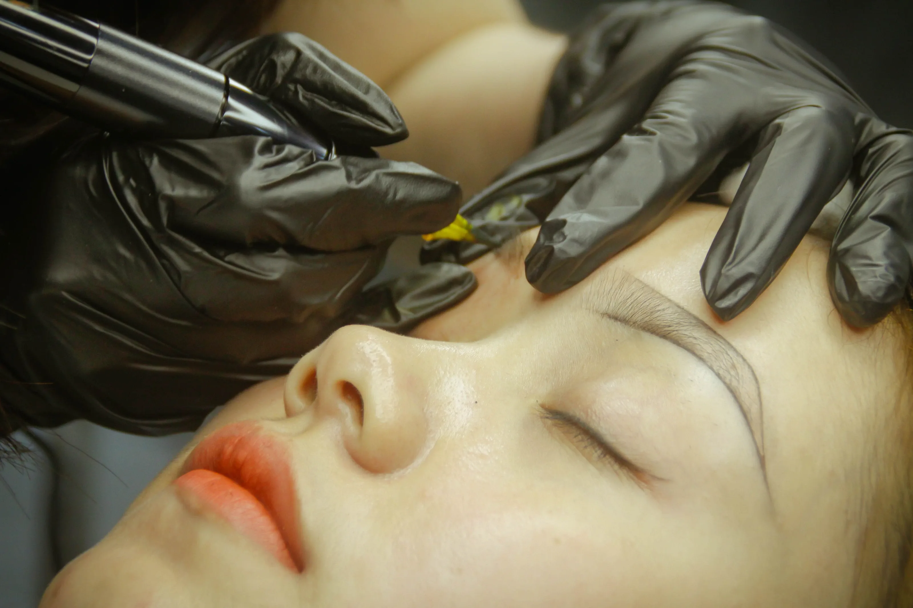 Micropigmentation pen working on a client's eyebrow while gloved hands hold the skin taut