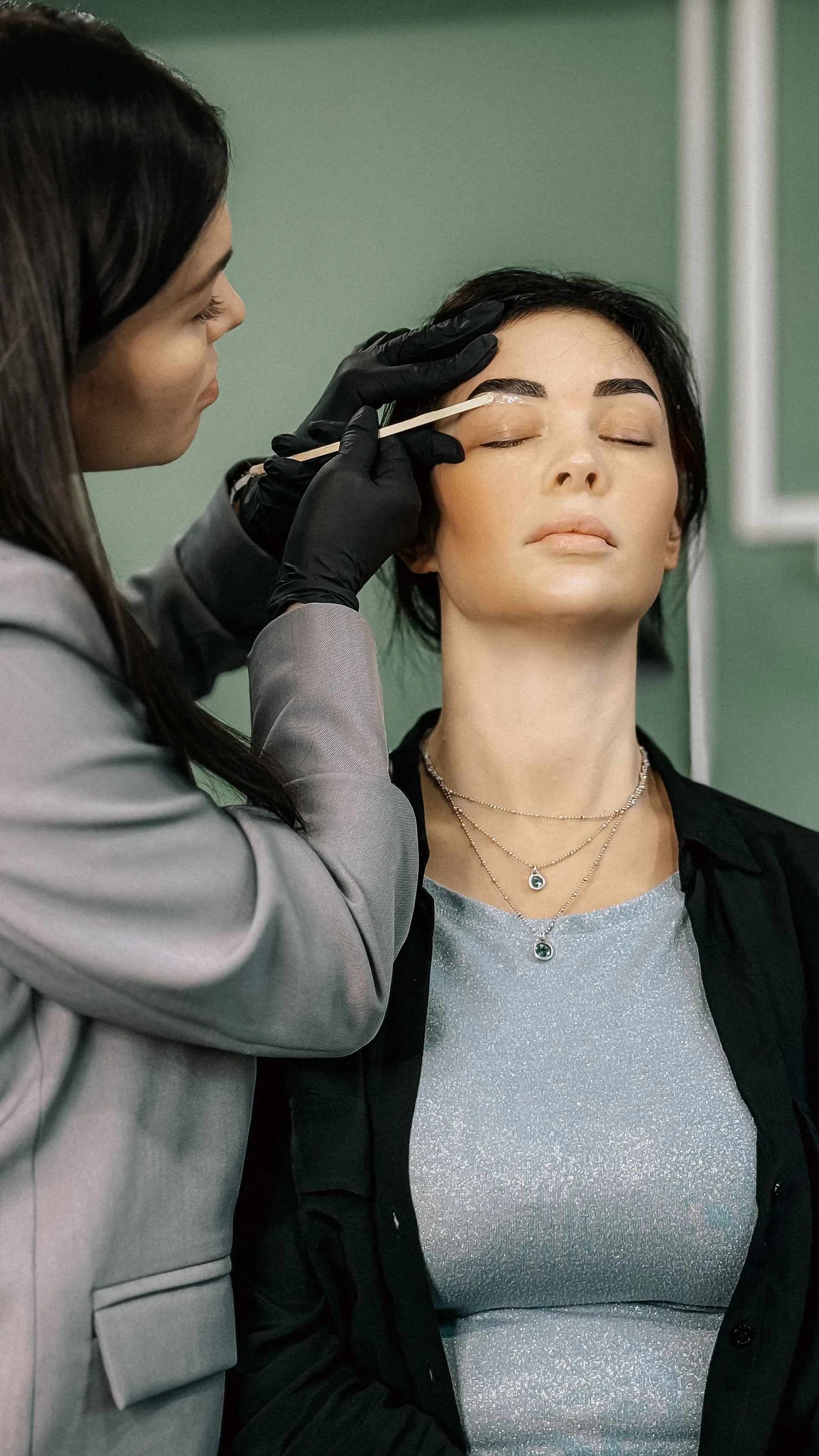 Technician in black gloves treating a client's eyebrow in a bright treatment room
